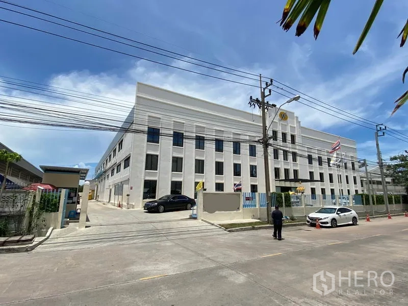 โรงงาน ให้เช่าหรือขายในBang Sao Thong, Samut Prakan - มุมมองจากถนนของโรงงานสมัยใหม่หลายชั้น มีกำแพงรั้ว ทางเข้า ที่จอดรถ และสายไฟเหนือศีรษะ ในวันที่อากาศแจ่มใส