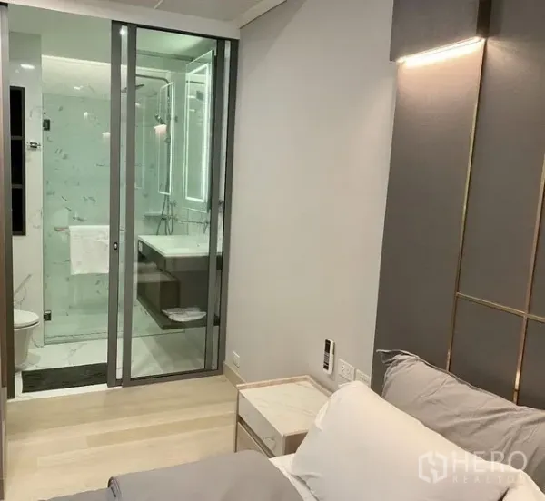 Condominium for rent in Khlong Toei, Bangkok - Bedroom view to glass-enclosed bathroom with illuminated mirror and light wood flooring.