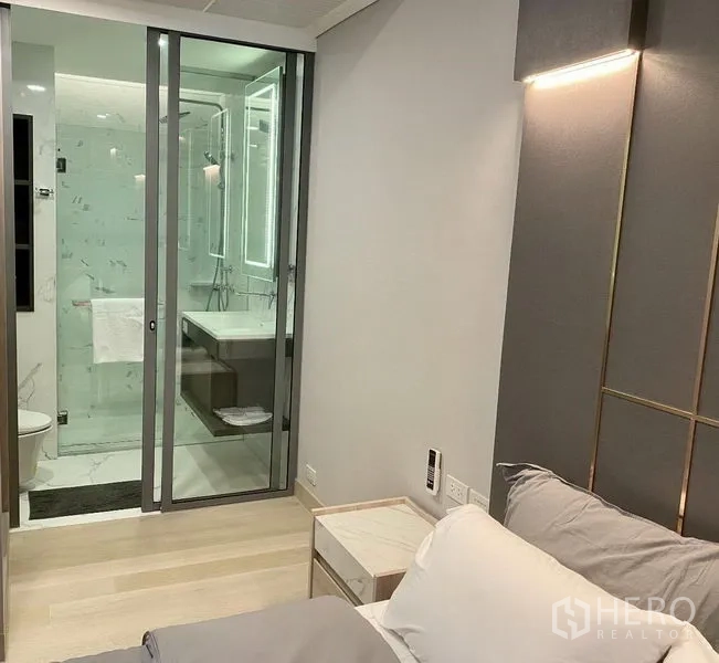 Condominium for rent in Khlong Toei, Bangkok - Bedroom view to glass-enclosed bathroom with illuminated mirror and light wood flooring.