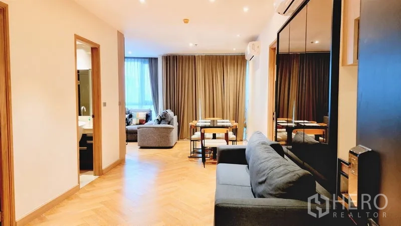 Condominium for sale in Watthana, Bangkok - Open living–dining space with sofa, dining table, mirrored storage and full-height curtains.