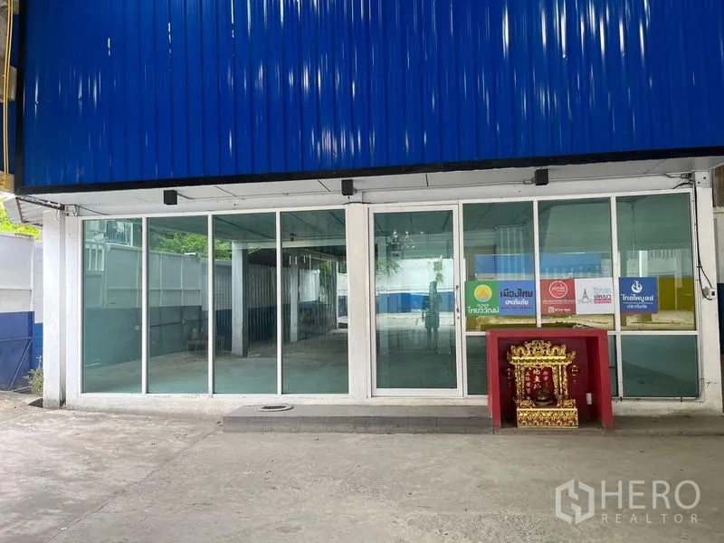 Warehouse for rent in Phra Khanong, Bangkok - Glass-fronted office entrance beneath blue metal cladding in the yard.