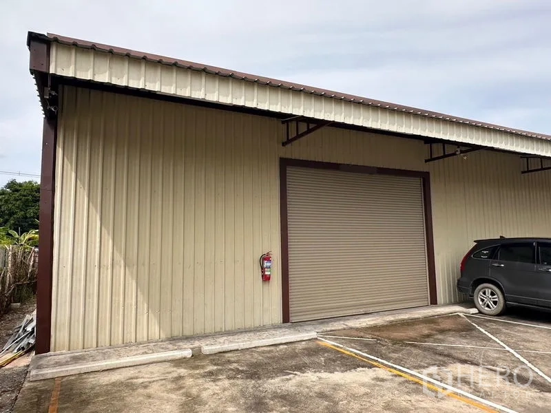 Warehouse for rent in Mueang Nonthaburi, Nonthaburi - Exterior of warehouse with closed roller shutter door, metal siding, and on-site parking spaces with a car.
