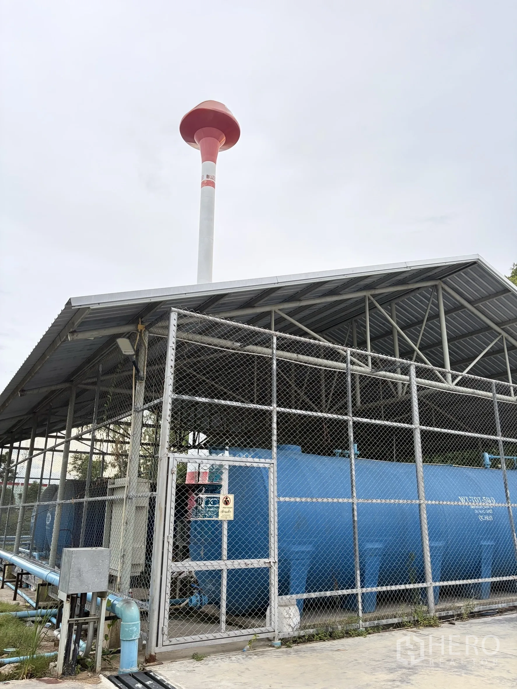 Factory for sale in Mueang Nakhon Pathom, Nakhon Pathom - Fenced utility zone housing large blue water tanks beneath a roof and water tower.