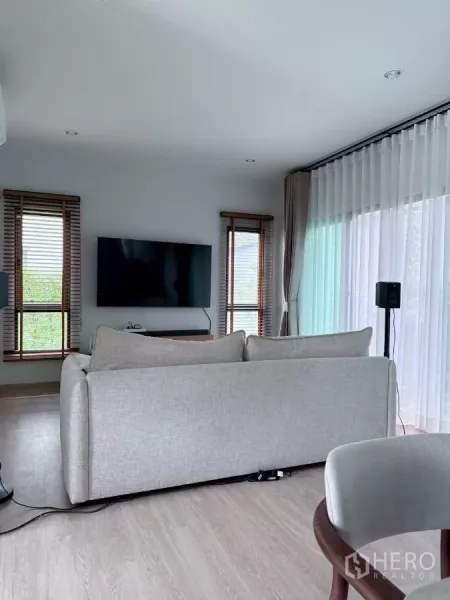 บ้าน ให้เช่าในLat Krabang, Bangkok - ห้องนั่งเล่นสว่างด้วยโซฟาสีเทา ทีวีติดผนัง และผ้าม่านโปร่ง