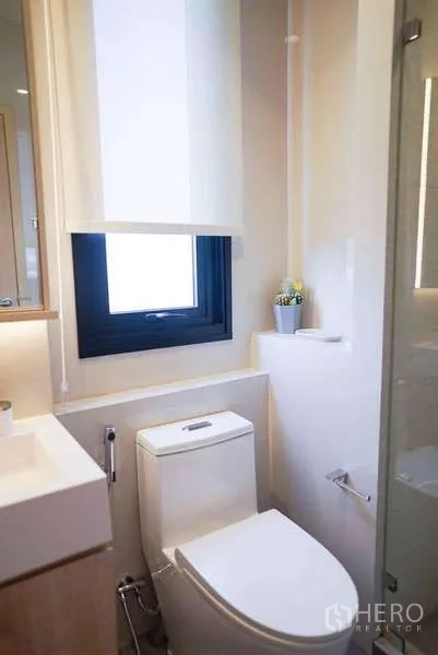 Condominium for rent or sale in Watthana, Bangkok - Compact bathroom with window, vanity, and white toilet beside a glass shower enclosure.