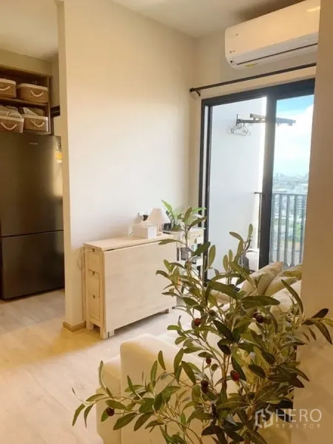 Condominium for sale in Suan Luang, Bangkok - Bright living room with cream sofa, light wood console, air conditioner and sliding glass door to a city-view balcony.