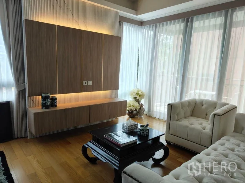 Condominium for rent or sale in Pathum Wan, Bangkok - Living area with illuminated built-in cabinet, tufted armchairs, coffee table, and wraparound windows.