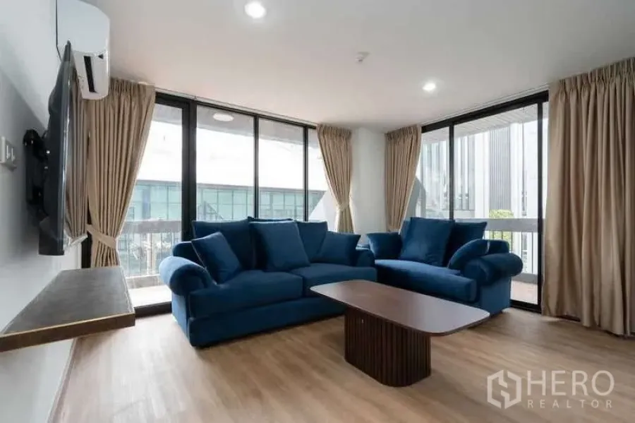 Apartment for rent in Khlong Toei, Bangkok - Corner living room with blue sofa set, wall-mounted TV and floor-to-ceiling windows.