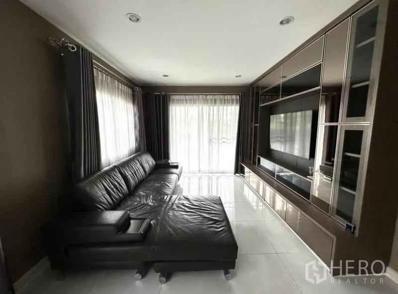 Detached House for rent in Suan Luang, Bangkok - Bright living room with black leather sectional sofa, built-in TV wall and floor-to-ceiling curtains.