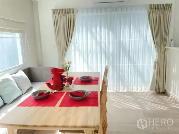 联排别墅 出租在Prawet, Bangkok - 餐桌靠窗摆放，红色餐垫与纱帘营造温馨用餐角。