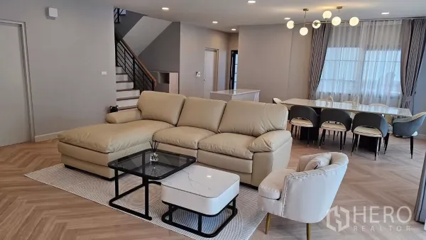 Detached House for rent or sale in Bang Phli, Samut Prakan - Cozy living room with beige L-shaped sofa, glass coffee table, staircase and eight-seat dining table on chevron flooring.