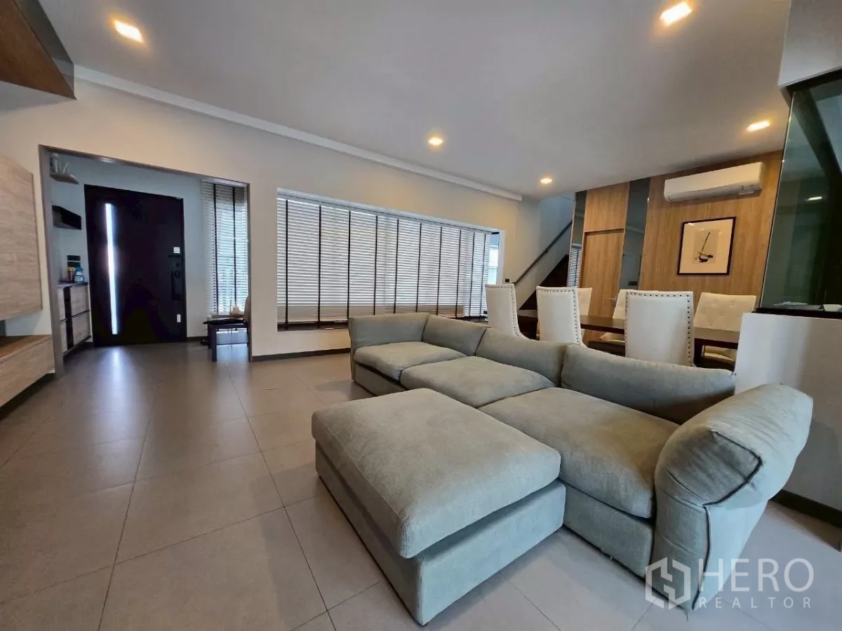 Townhouse for rent in Bang Phli, Samut Prakan - Spacious living area with sectional sofa, floor-to-ceiling blinds, recessed lighting, and the entryway in view.