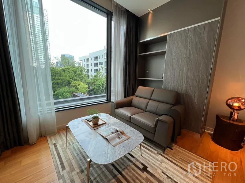 Condominium for rent in Pathum Wan, Bangkok - Cozy seating area with grey sofa, marble coffee table, built‑in shelves and large window with city and greenery view