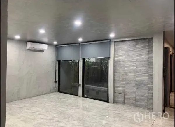 Detached House for rent or sale in Wang Thonglang, Bangkok - Ground-floor living area with large sliding windows, roller blinds, gray feature wall and recessed lights.