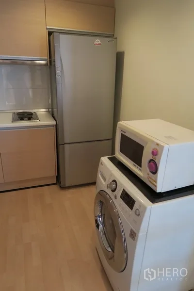 Condominium for rent in Huai Khwang, Bangkok - Compact kitchen with tall fridge, built‑in hob, light wood cabinetry, microwave on top of a front‑load washing machine.