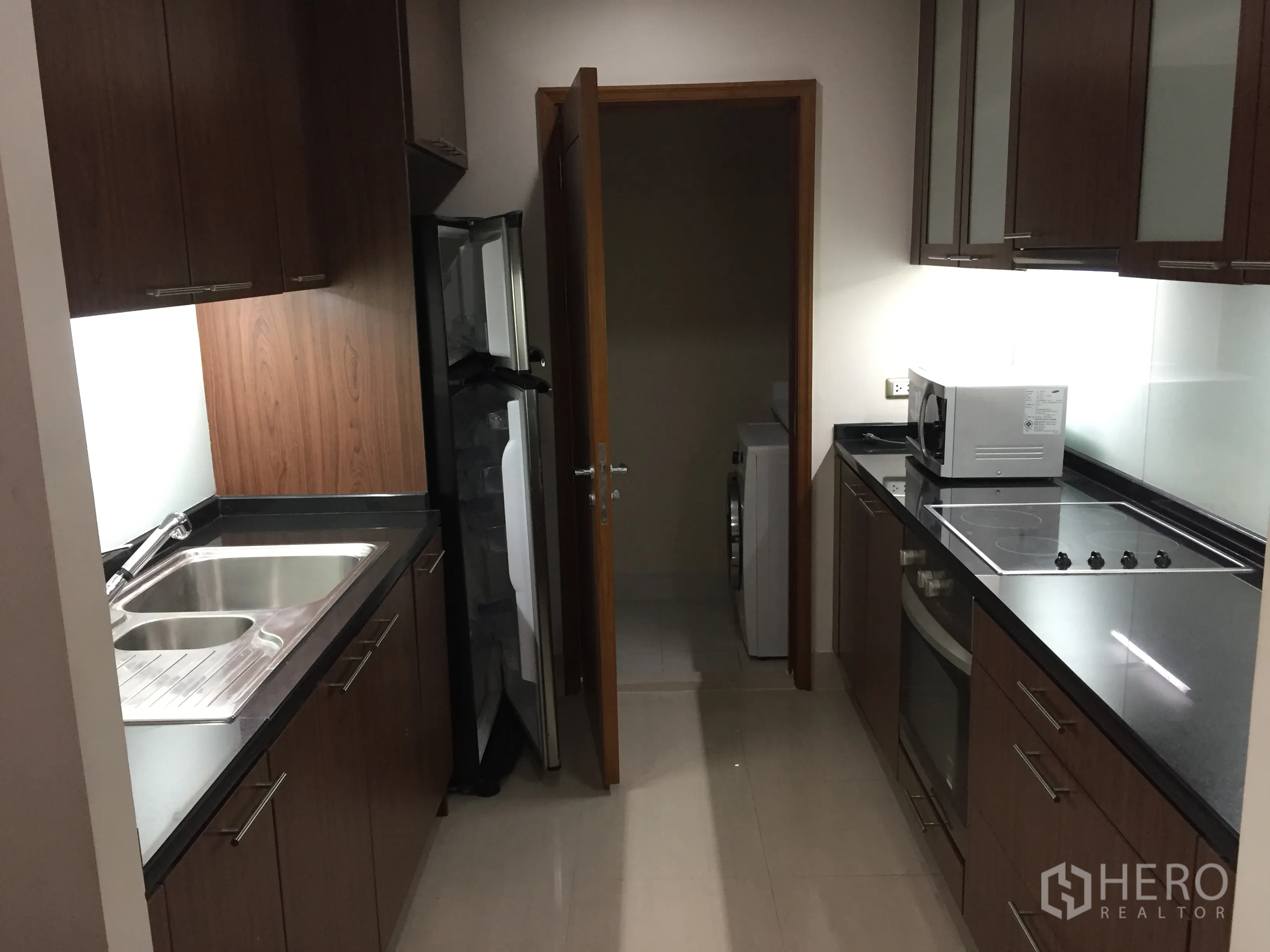 Condominium for rent in Sathon, Bangkok - Galley kitchen with wood cabinets, black stone counters, electric hob and oven, fridge, and a utility area with washing machine.