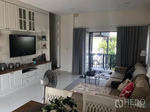 Townhouse for rent or sale in Saphan Sung, Bangkok - Living room with custom media cabinet opening to a balcony through sliding glass doors with greenery view.