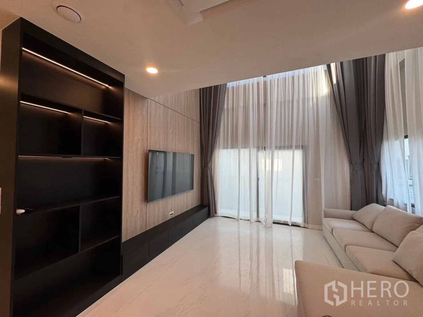 Detached House for rent in Bang Phli, Samut Prakan - Living room with floor-to-ceiling sheer curtains, TV, and dark shelving.