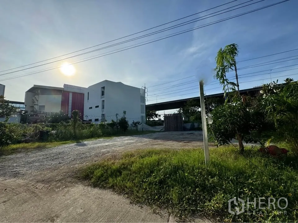 Land for rent in Bang Na, Bangkok - Cleared gravel lot with paved entrance beside a low-rise building and an elevated roadway.