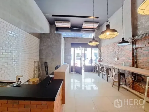 零售空间 出租在Watthana, Bangkok - 工业风商铺内景，高挑层空间，砖与瓷砖墙面，吊灯、长吧台与临街玻璃门面。