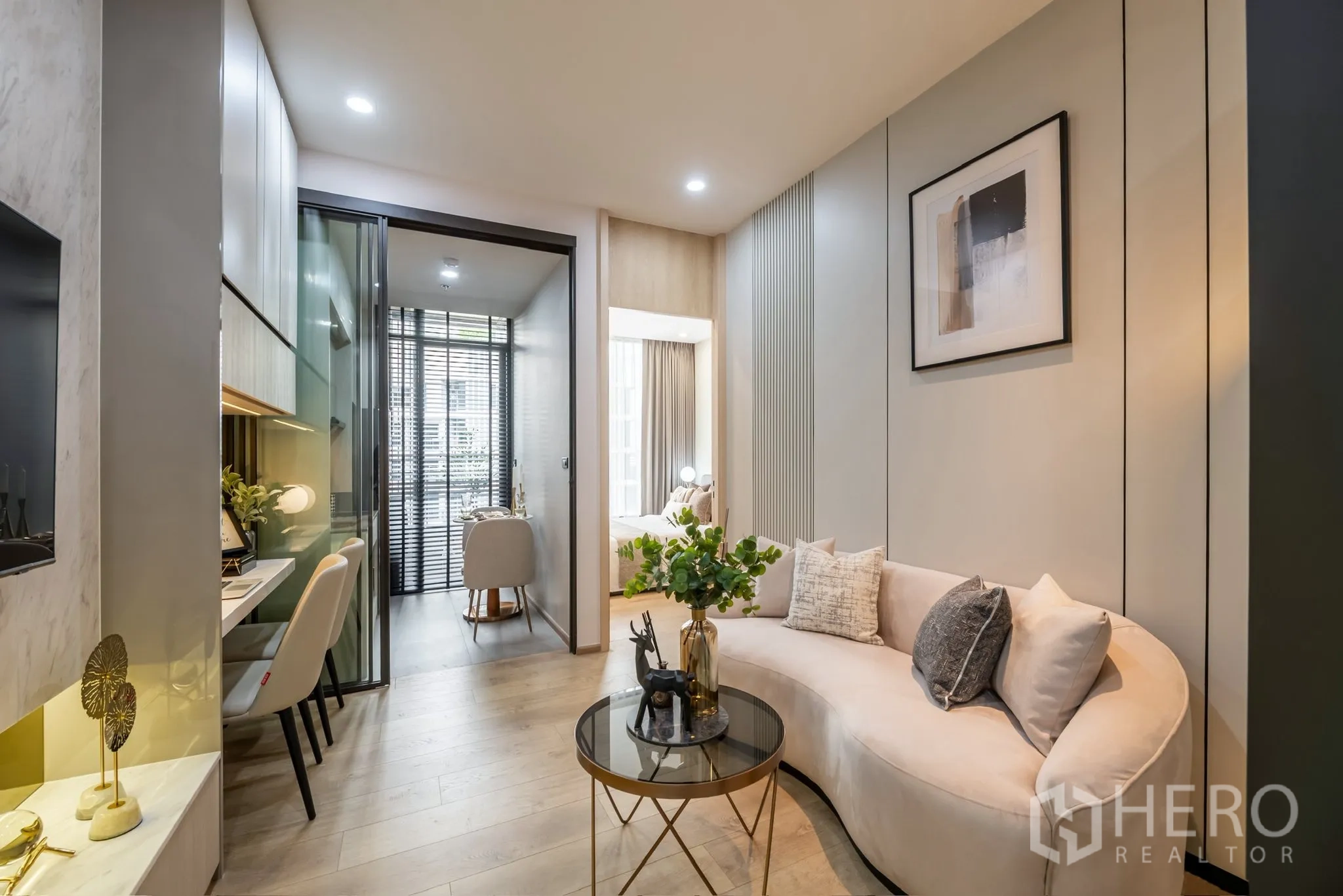 Condominium for rent in Watthana, Bangkok - Bright living room with a curved sofa, round glass coffee table, built-in study desk, sliding glass partition, and dining nook by tall windows.