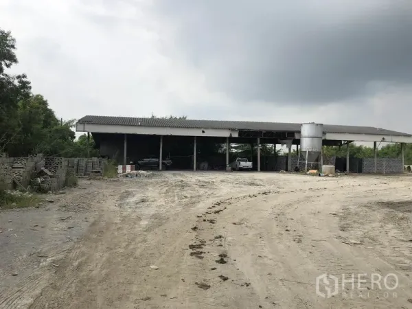 工厂 出租在Phra Nakhon Si Ayutthaya - 开放式车棚配有立式筒仓和土路车道，周围是树木与堆放材料。