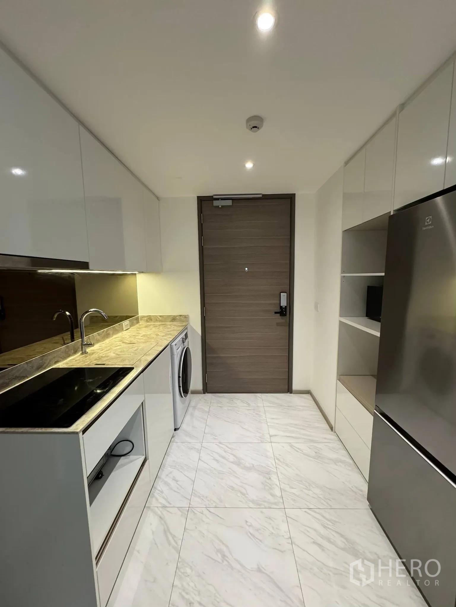 Condominium for rent in Khlong Toei, Bangkok - Sleek galley kitchen with stone-look countertop, cooktop, washing machine and large fridge.