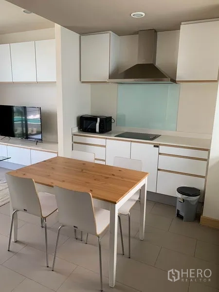 Condominium for rent in Watthana, Bangkok - Compact kitchen and dining area with TV unit visible, white cabinets and a stainless range hood.