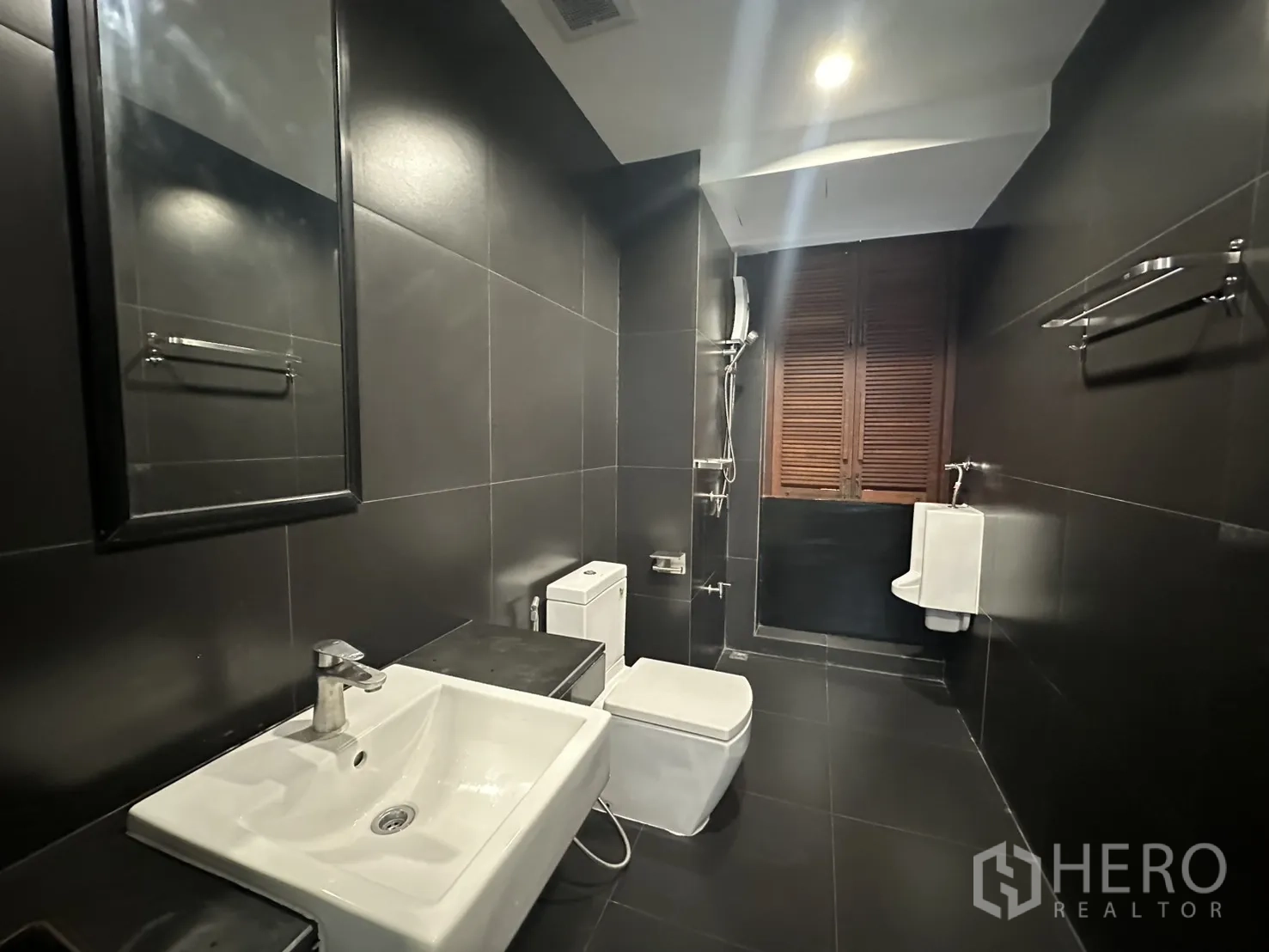 Apartment for rent in Pathum Wan, Bangkok - Modern bathroom with dark tiles, sink, toilet, urinal, and a shower area.