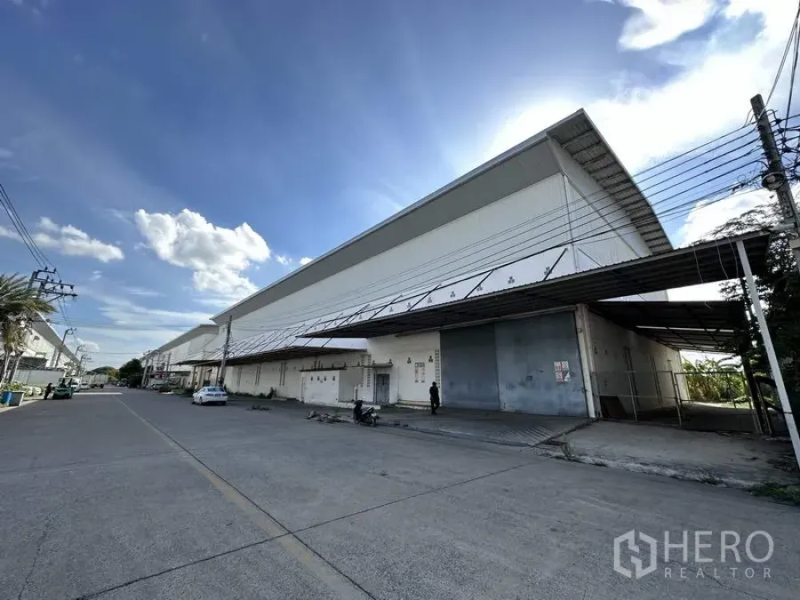 工厂 出租在Bang Bua Thong, Nonthaburi - 沿街长立面的工厂，金属大门与雨棚映衬蓝天。
