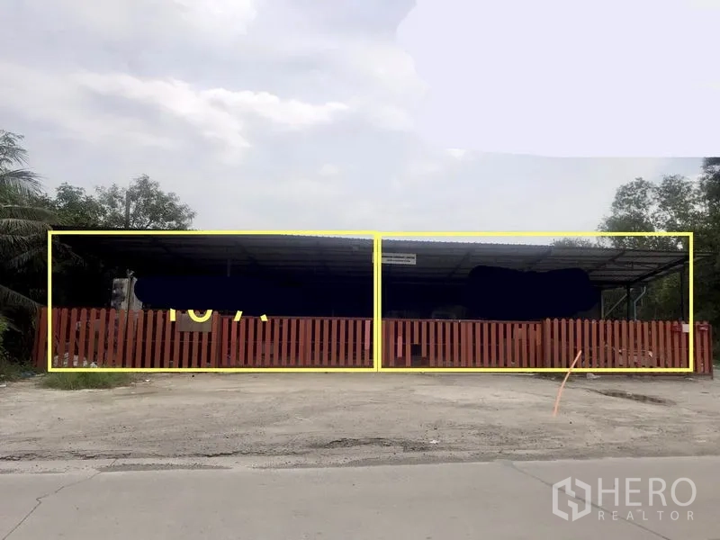Warehouse for rent in Pathum Thani - Front view of a gated warehouse with a long steel roof and red slat fence behind a concrete apron.