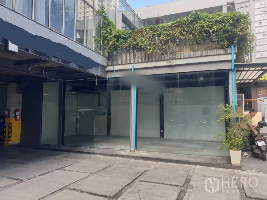 零售空间 出租在Khlong Toei, Bangkok - 一层临街商铺，整面落地玻璃门面，前方混凝土空地，上方阳台有绿植。