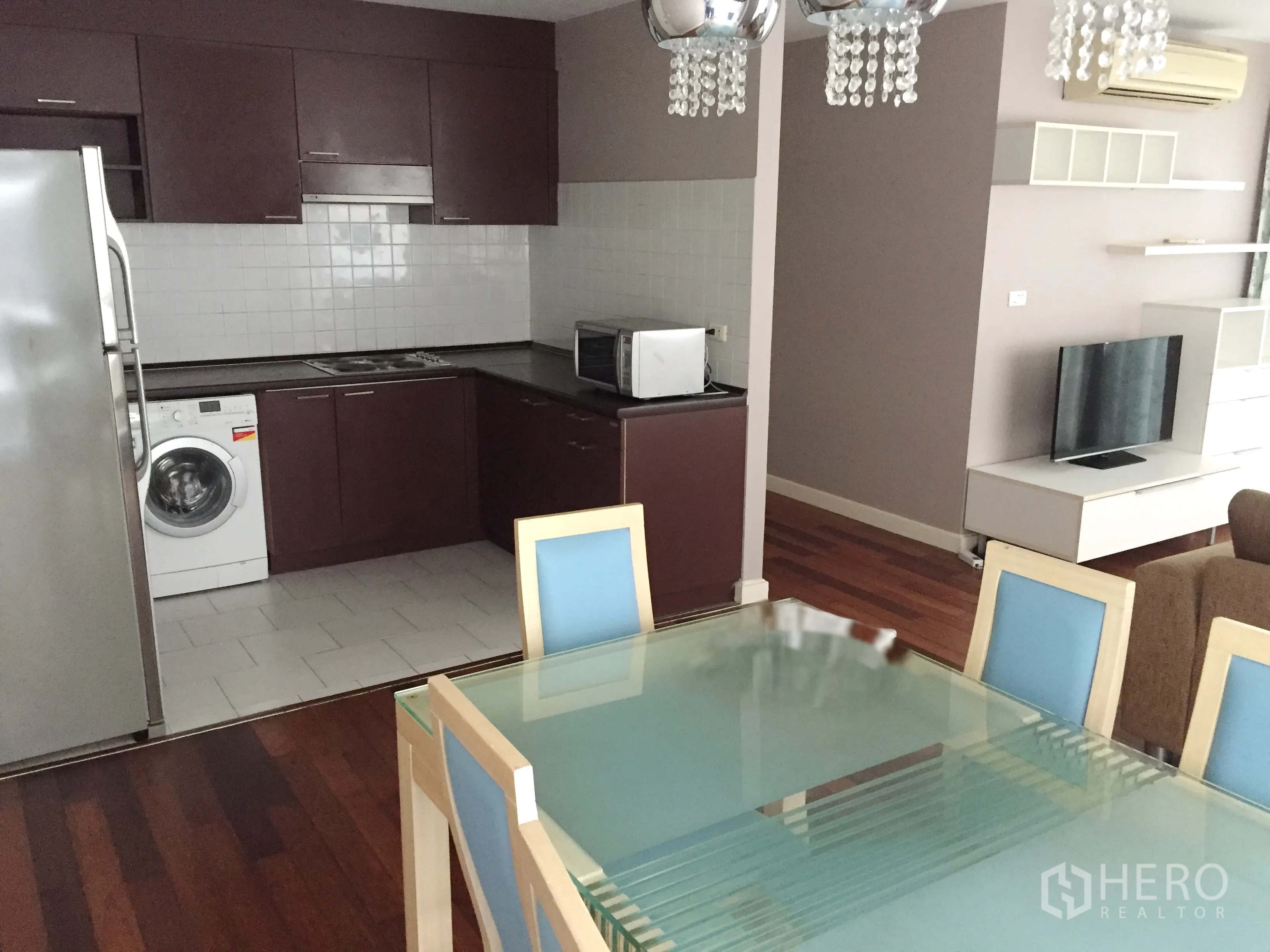 คอนโด ให้เช่าในWatthana, Bangkok - โต๊ะทานอาหารกระจกสำหรับหกที่นั่ง มองไปยังครัวรูปตัวแอลพร้อมเครื่องซักผ้าและชั้นวางทีวี