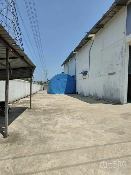 工厂 出售在Sai Noi, Nonthaburi - 厂房侧大面积混凝土场地，蓝色帐篷与架空电线。