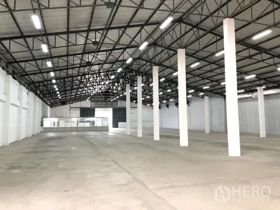 货栈 出租在Nonthaburi - 仓库内部空间宽敞，钢结构高屋顶、LED照明、白色立柱与混凝土地面，后方可见玻璃隔断办公室。