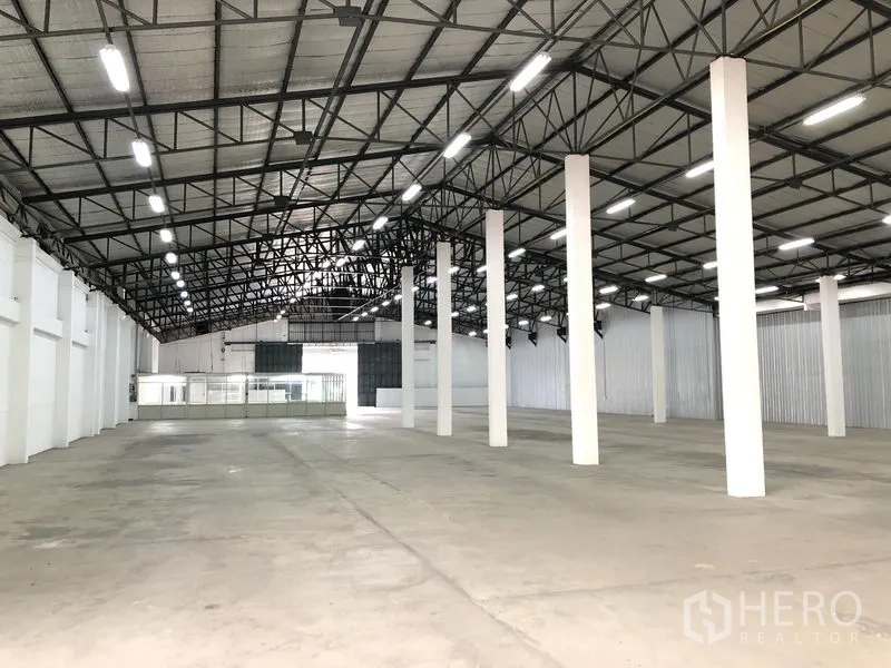 货栈 出租在Nonthaburi - 仓库内部空间宽敞，钢结构高屋顶、LED照明、白色立柱与混凝土地面，后方可见玻璃隔断办公室。