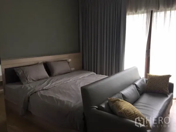 公寓 出租或出售在Bangkok - 温馨卧室，双人床，灰色窗帘，通往阳台的推拉落地门，床尾设有沙发。