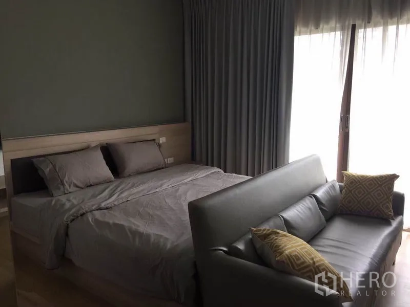 公寓 出租或出售在Bangkok - 温馨卧室，双人床，灰色窗帘，通往阳台的推拉落地门，床尾设有沙发。