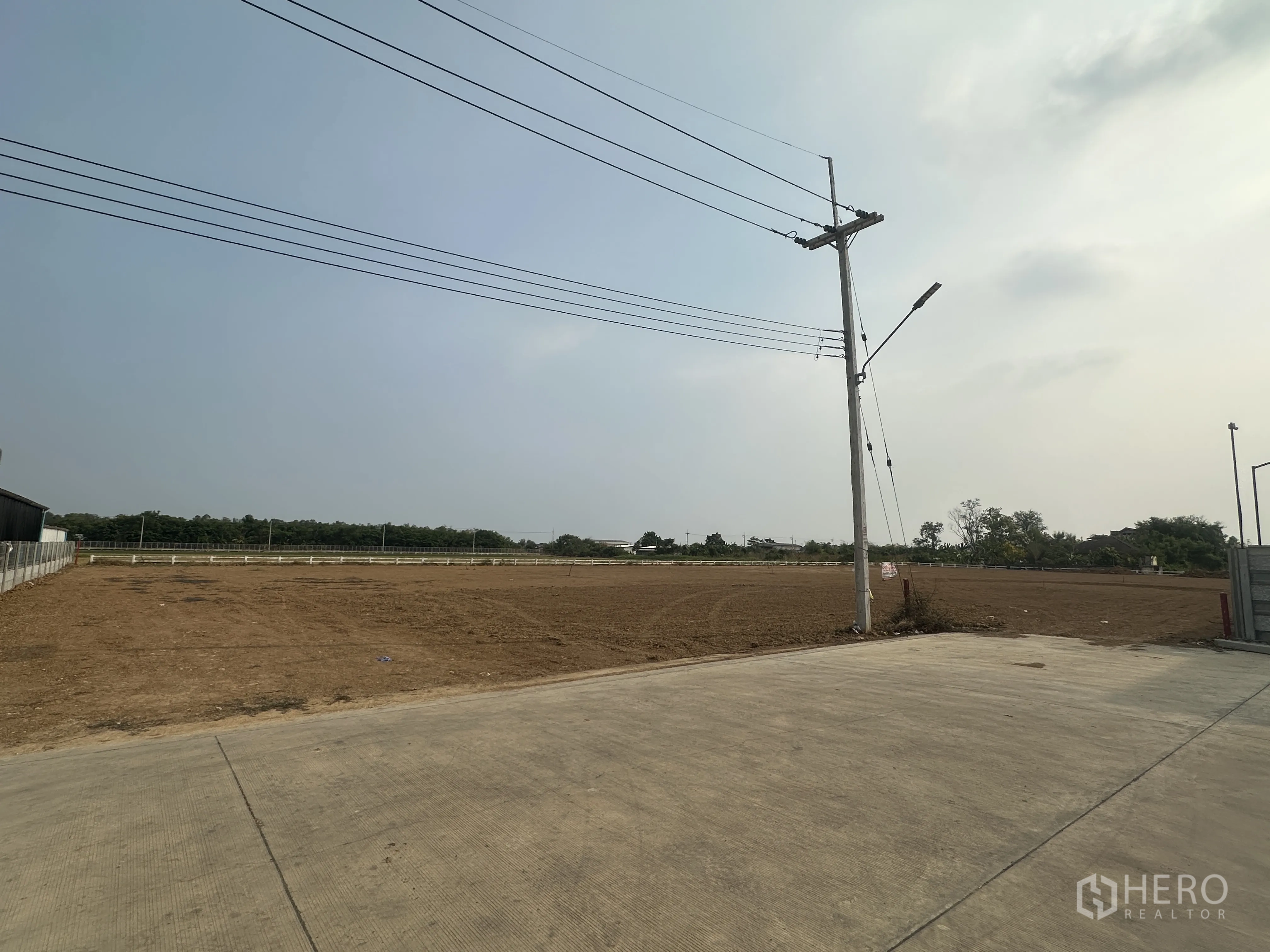 土地 for sale in Lam Luk Ka, Pathum Thani - 清理后的平整地块，邻接宽阔混凝土道路，设有电线杆与架空电缆，周边视野开阔。