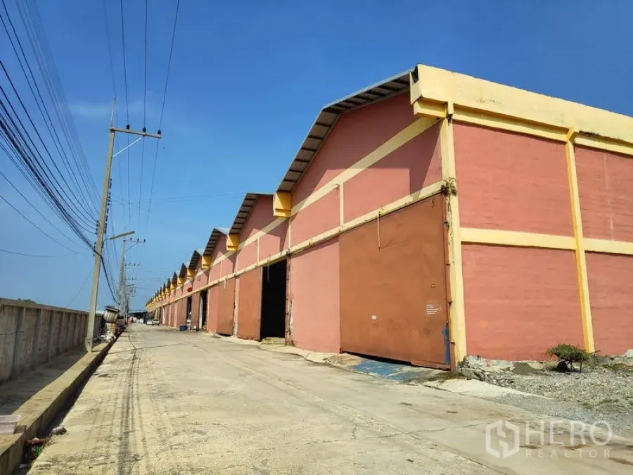 工厂 出租在Sai Noi, Nonthaburi - 连排仓库式工厂沿混凝土道路分布，配金属大门与道路电线杆。