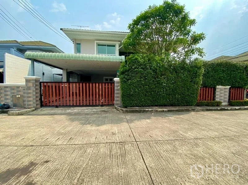 Detached House for sale in Phasi Charoen, Bangkok - Front exterior of the detached house with tall hedges, red wooden gate, and covered carport.