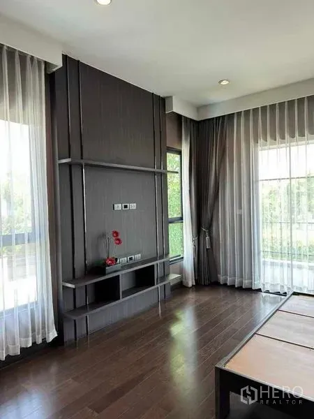 Detached House for sale in Prawet, Bangkok - Corner room with dark wood flooring, built-in TV wall and sheer curtain windows.