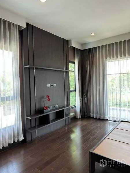 Detached House for sale in Prawet, Bangkok - Corner room with dark wood flooring, built-in TV wall and sheer curtain windows.
