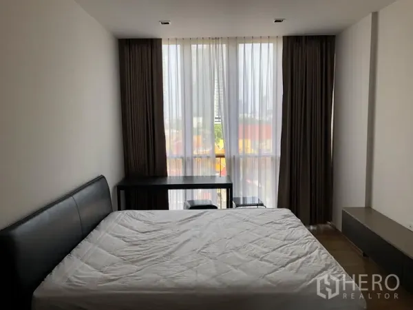 Condominium for rent in Watthana, Bangkok - Primary bedroom featuring large windows, blackout drapes, and a study desk overlooking the city.