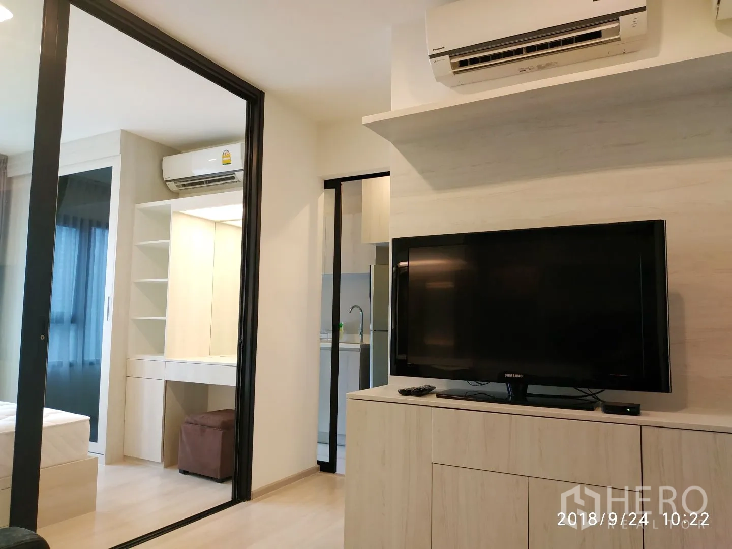 Condominium for rent in Huai Khwang, Bangkok - Living area with TV console and air conditioning, connected to kitchen and bedroom by glass doors.