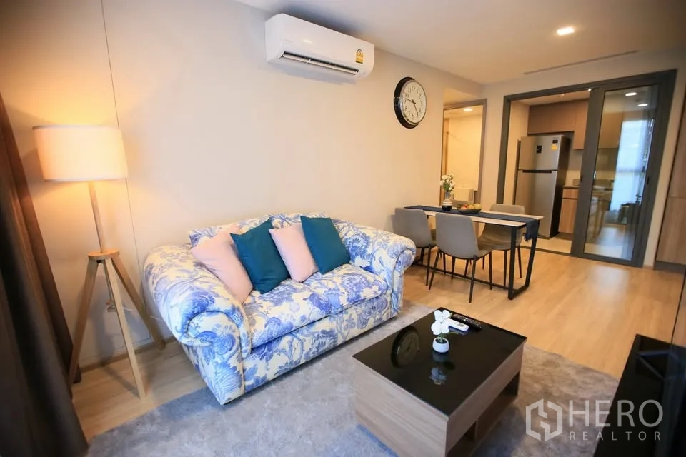 Condominium for rent in Watthana, Bangkok - Cozy living room with floral sofa, dining for four and kitchen behind sliding glass.
