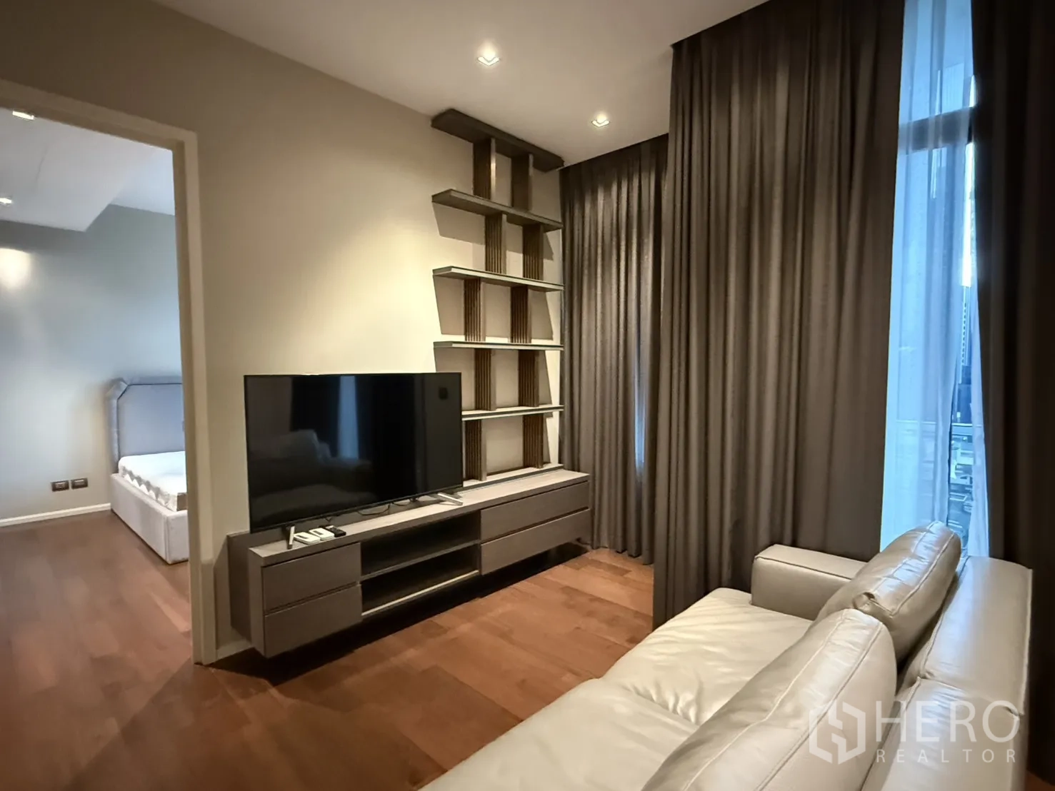 Condominium for rent in Watthana, Bangkok - Contemporary living room with leather sofa, TV console, built-in shelving, wood floors and tall curtains, with a view into the bedroom.