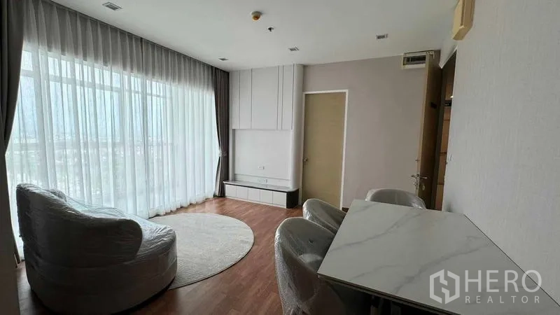 คอนโด ให้เช่าในBang Na, Bangkok - มุมนั่งเล่นมีผนังกระจกบานใหญ่ พรมทรงกลม ตู้วางทีวีบิวท์อิน และโต๊ะกินข้าวท็อปหินอ่อนบนพื้นไม้