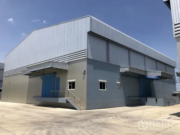 Warehouse for rent in Lat Lum Kaeo, Pathum Thani - Modern steel-clad warehouse exterior with wide concrete yard and covered loading docks under a clear sky.