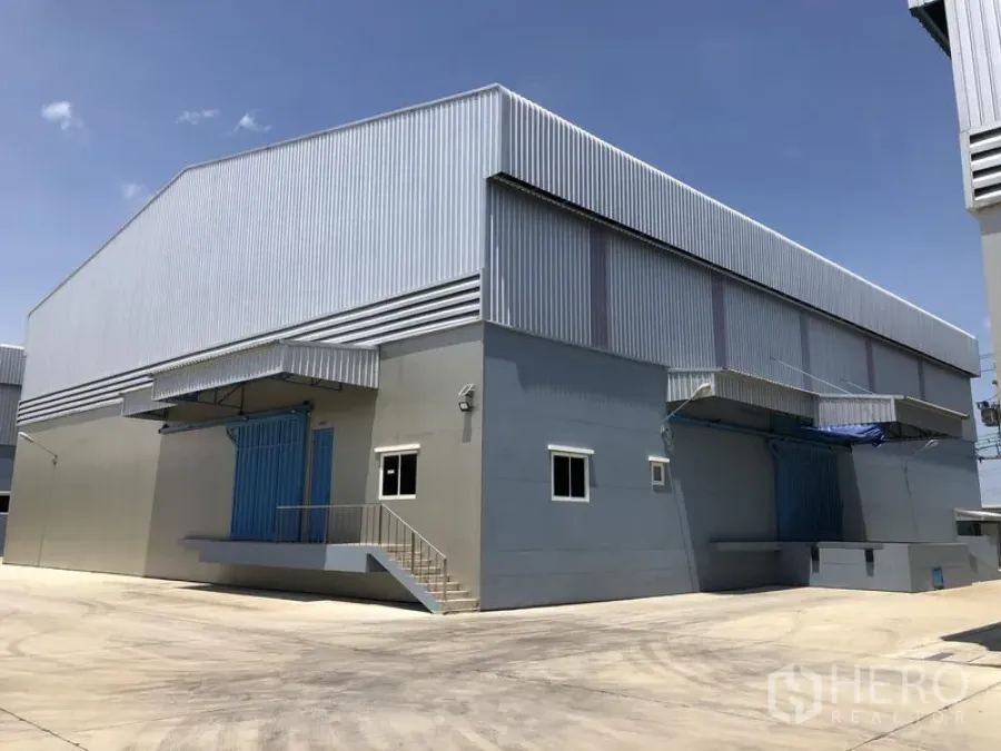 Warehouse for rent in Lat Lum Kaeo, Pathum Thani - Modern steel-clad warehouse exterior with wide concrete yard and covered loading docks under a clear sky.